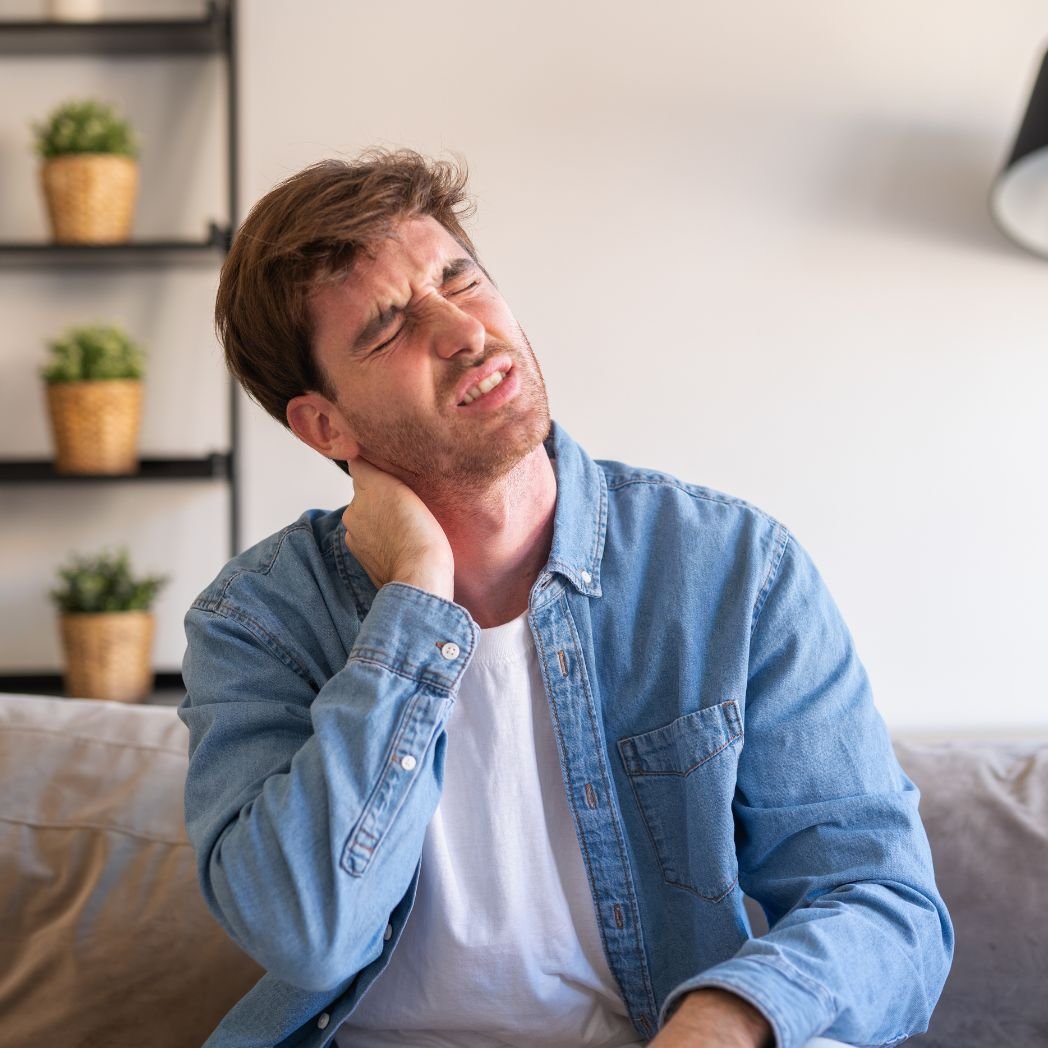 “Person sitting indoors holding the side of their neck with a painful expression, illustrating joint stiffness, muscle tension, or neck discomfort in a home setting.”