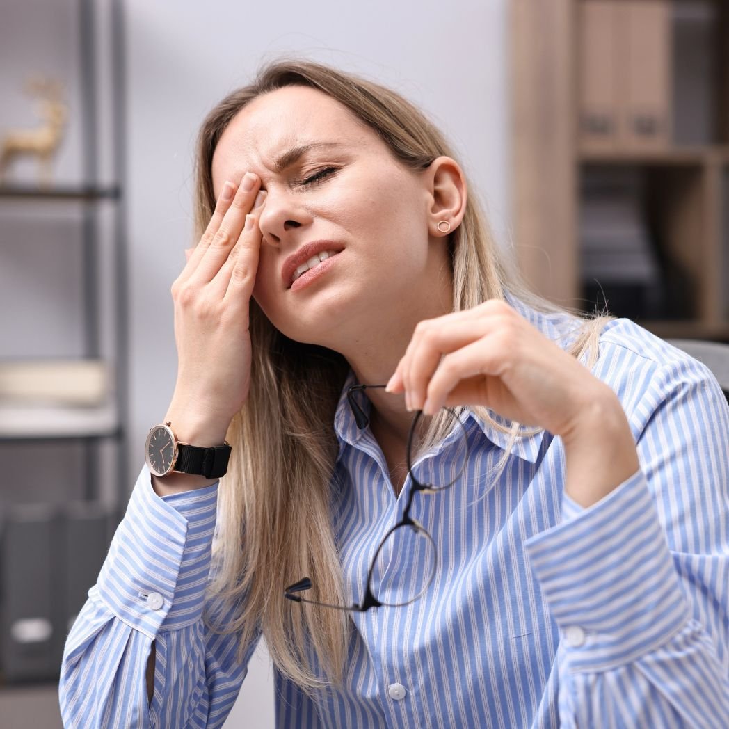 Woman experiencing persistent fatigue and headache symptoms related to immune dysregulation while sitting at office desk.